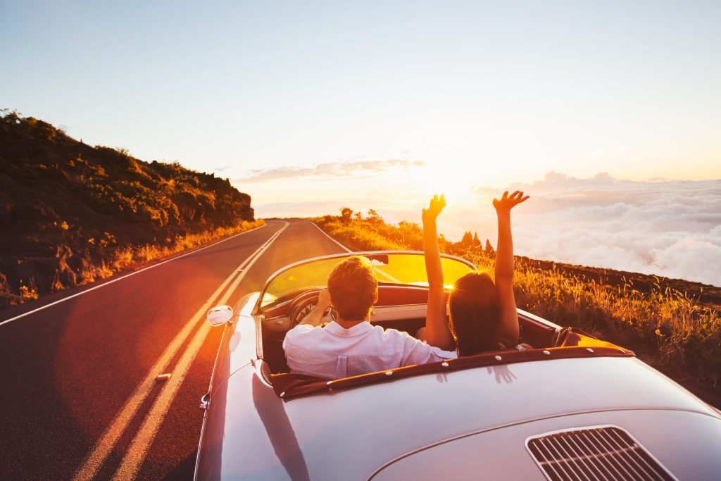 Summer Road Trip Maintenance has been done causing a couple in a convertible vehicle to enjoy the lakeside drive worry=free with hands lifted high in the air as the sun rises.