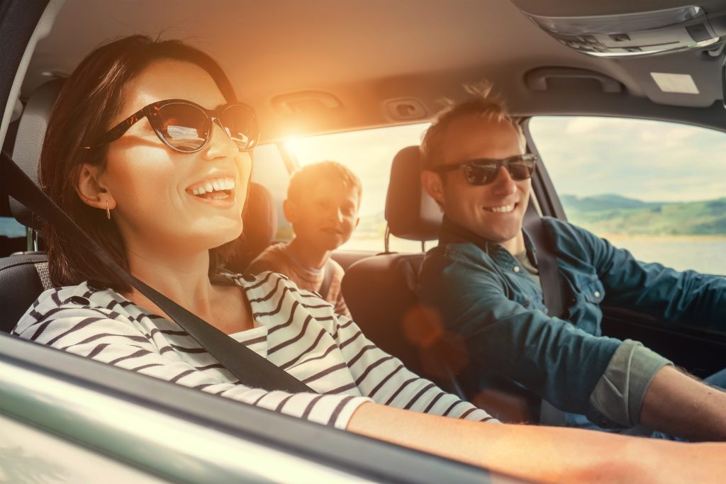Smiling parents and their enthusiastic son enjoy a GMC SUV Summer Road Trip as the sun shines through the back window of the vehicle in the countryside.